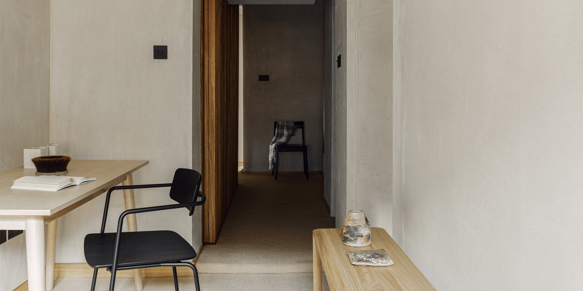 A minimalist hallway featuring a wooden table, a black chair, and a chair with a plaid throw, all against soft, neutral-toned walls.