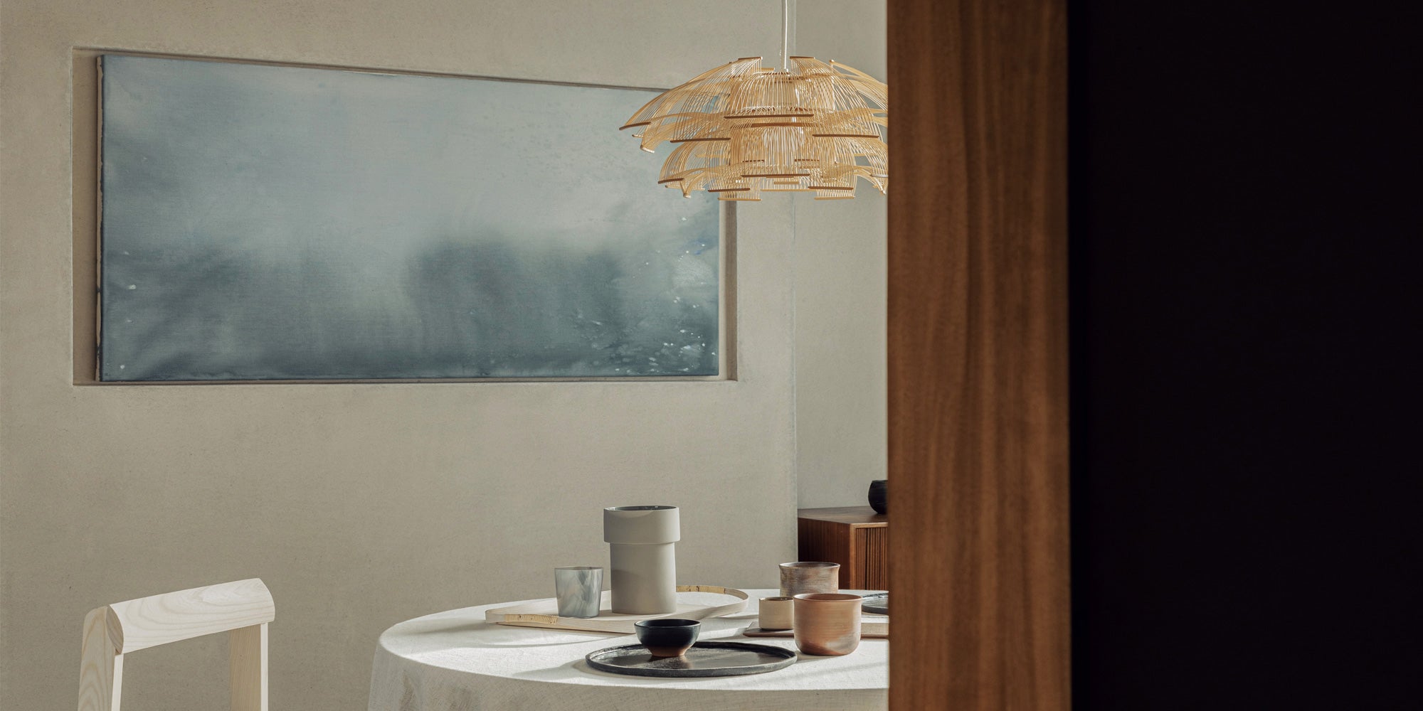 A modern dining space with a round table set with minimalist plates and cups, illuminated by a stylish bamboo pendant light.
