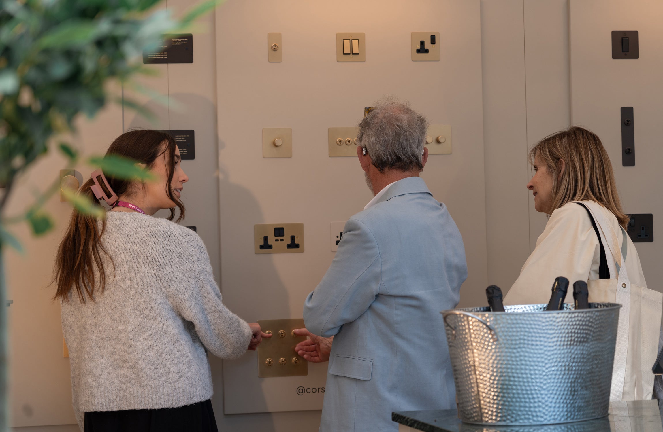 Three individuals examine various light switches and electrical outlets on a display wall, engaging in a design discussion.