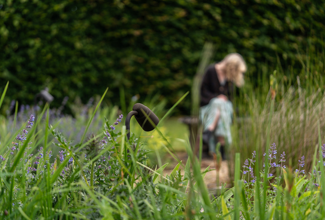 Out-of-focus person sat on a garden bench, with a bronze 'Ealing Path Spotlight' featuring in the foreground.