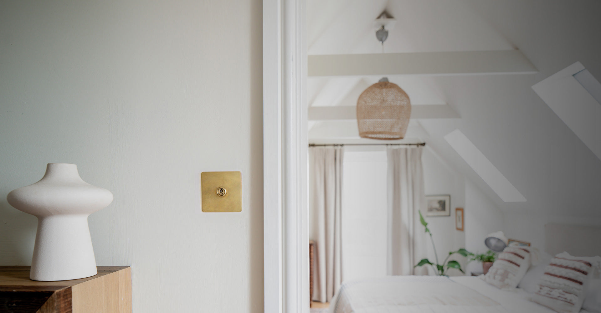A minimalistic interior featuring a white vase, brass light switch, and natural light, highlighting a stylish living space.