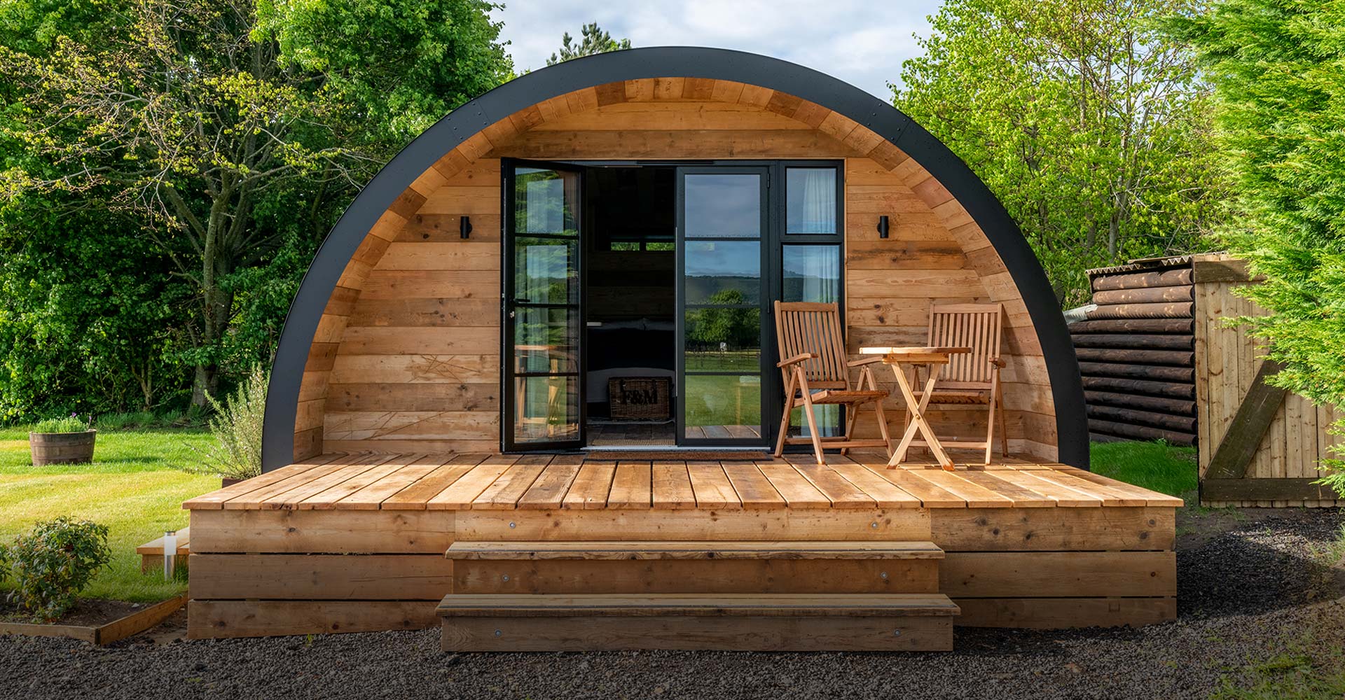 A modern wooden cabin with large glass doors, two outdoor chairs, and a table, surrounded by greenery in a sunny setting.
