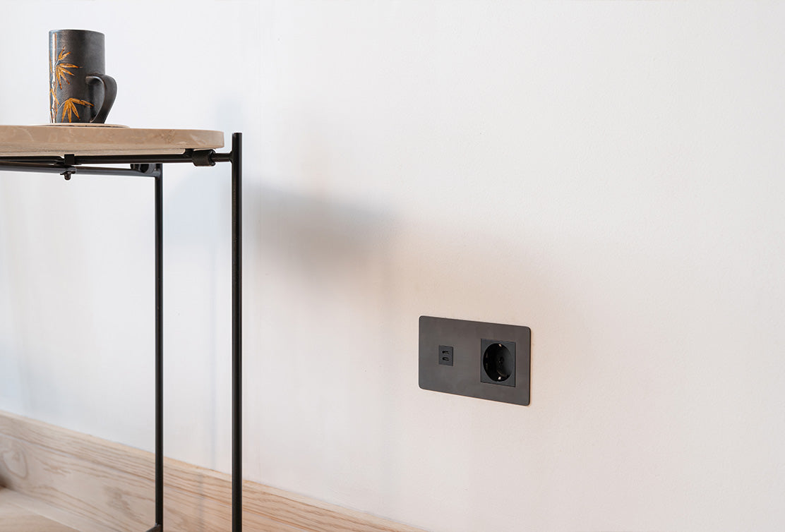 A minimalist black mug rests on a marble-topped table, next to a wall-mounted electrical outlet. Light-colored wood flooring below.