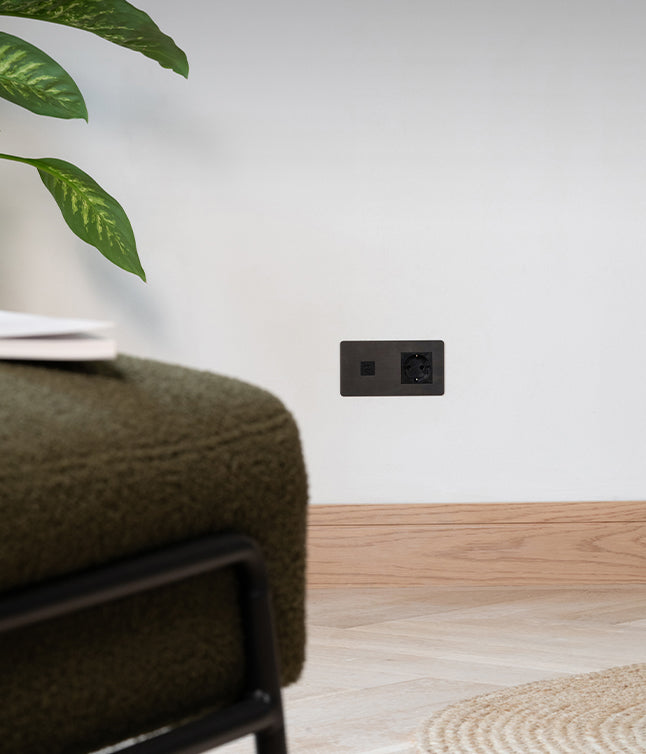 A stylish black power outlet embedded in a white wall, beside a green plant and a textured dark green ottoman.