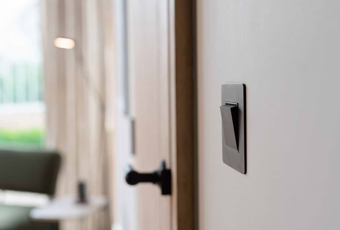 A close-up of a sleek black light switch mounted on a beige wall, with a wooden door and soft lighting in the background.