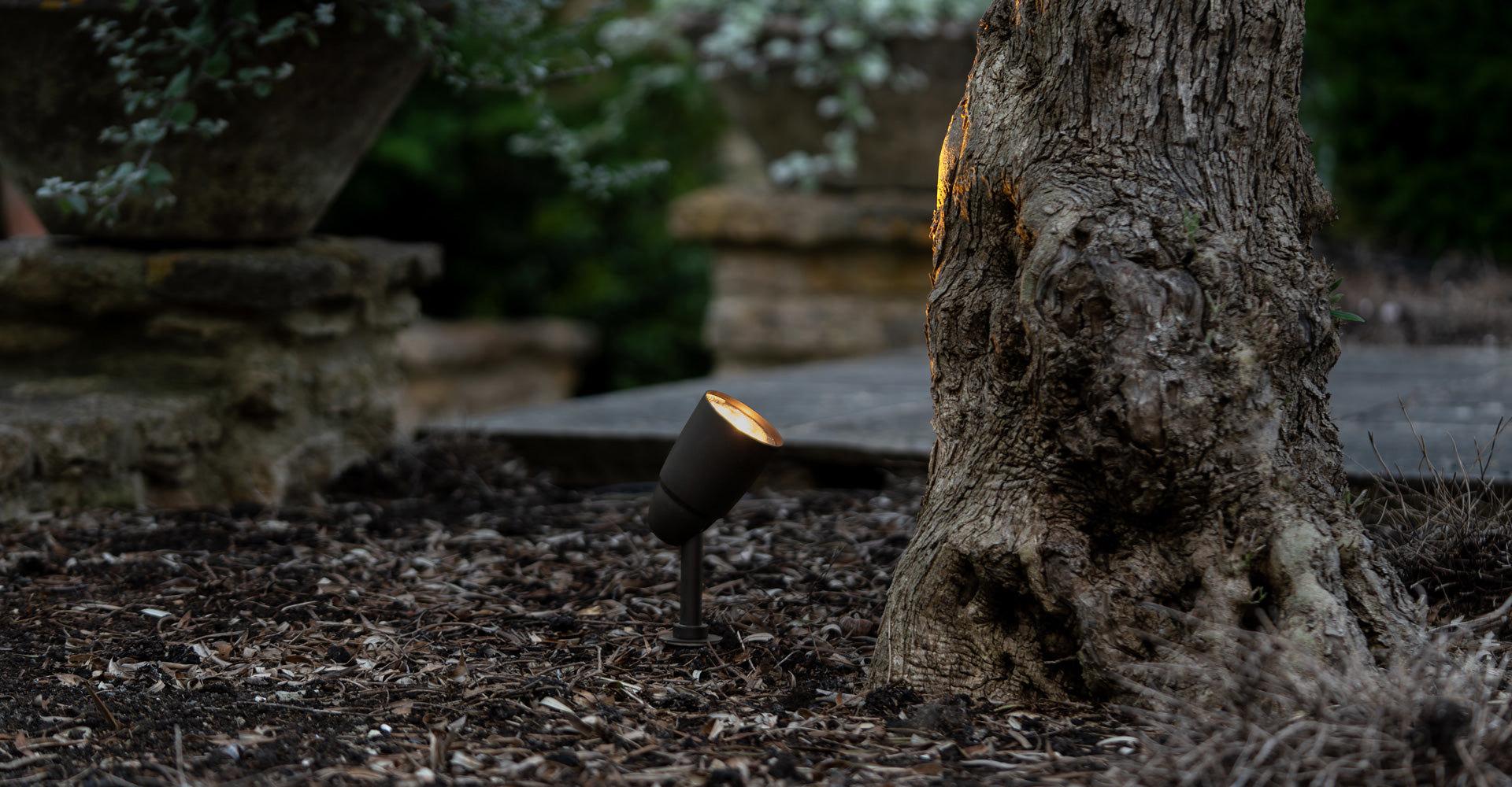 Standalone bronze 'Ealing Path Spotlight' illuminating a terracotta bench in front of a brown background. 