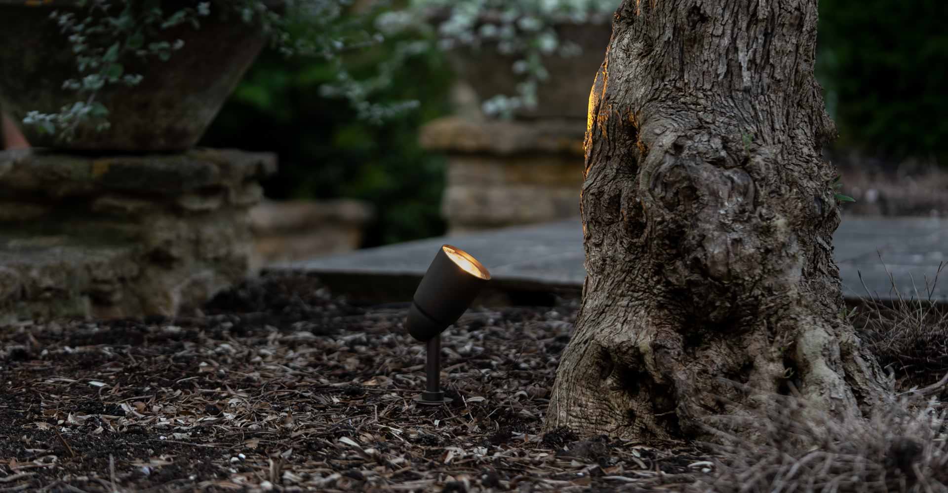 Standalone bronze 'Ealing Path Spotlight' illuminating a terracotta bench in front of a brown background.