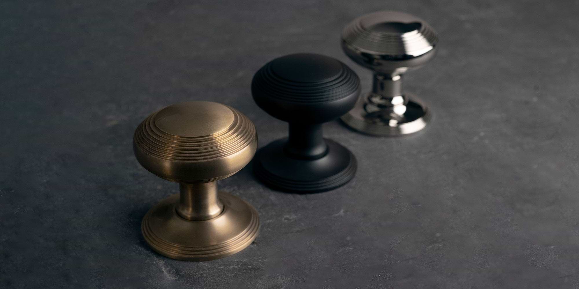 Three decorative cabinet knobs in different finishes: antique brass, matte bronze, and polished nickel, arranged on a dark surface.