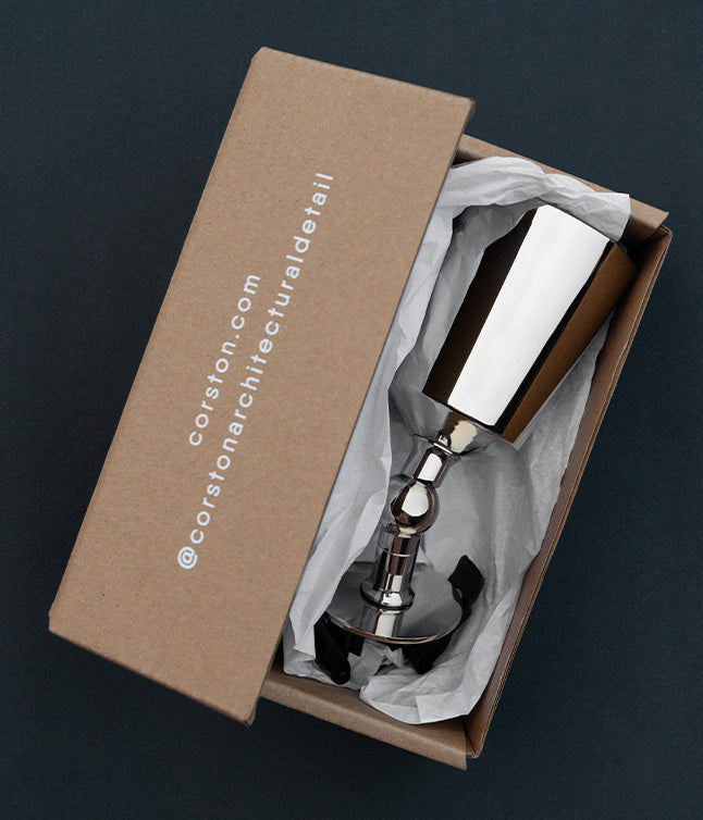 A polished nickel spotlight inside a recycled cardboard box, lined with white tissue paper, showcasing elegant packaging design.