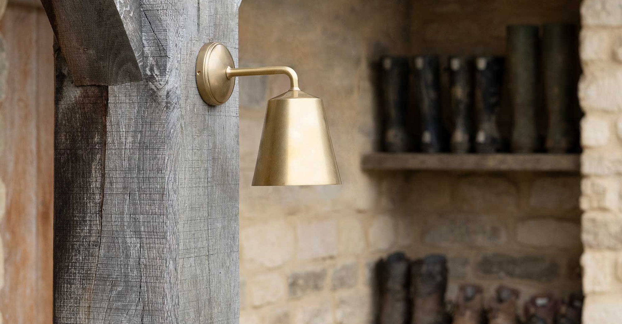 A rustic brass sconce illuminates a textured wooden beam, with earthy stone walls and blurred boots on a shelf in the background.
