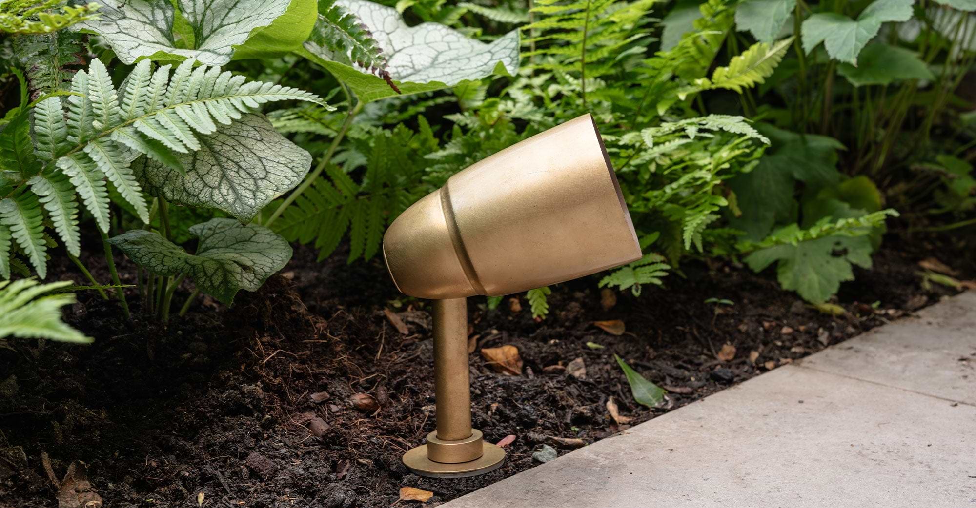 A golden garden spotlight positioned at an angle among lush green plants and dark soil, enhancing outdoor lighting aesthetics.