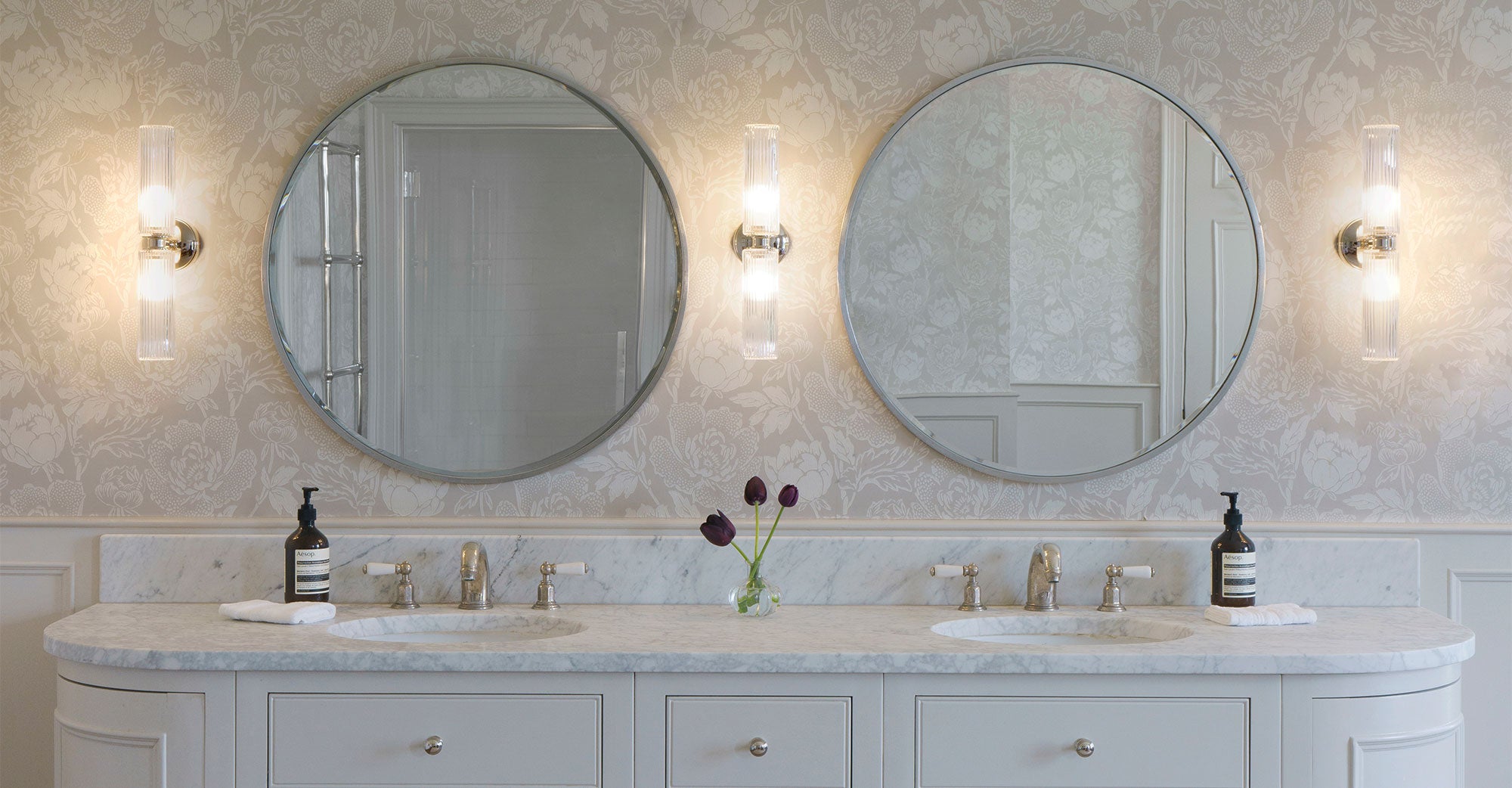 Elegant bathroom featuring dual round mirrors, marble countertop, modern lighting, and a vase with purple tulips against floral wallpaper.