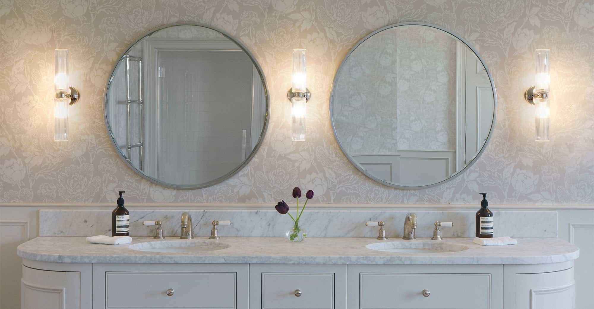 Elegant bathroom featuring dual round mirrors, marble countertop, modern lighting, and a vase with purple tulips against floral wallpaper.