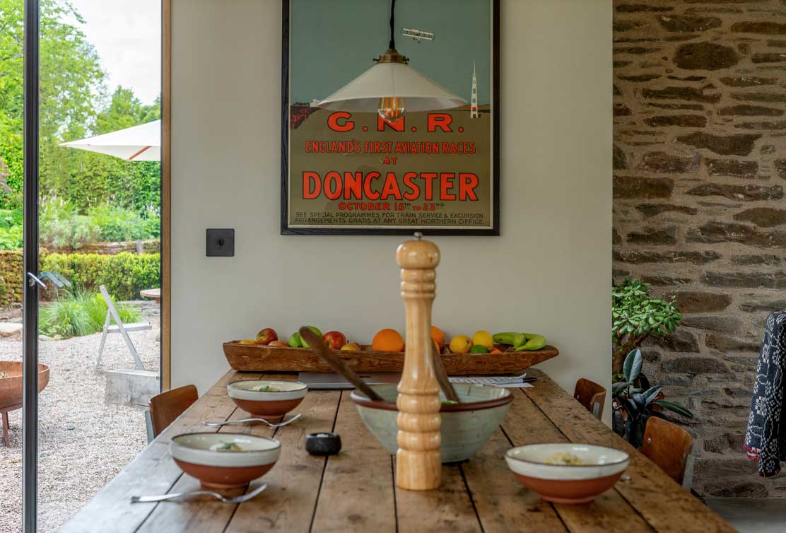 A rustic dining table set with bowls and a wooden pepper mill, framed by a vibrant aviation race poster and lush outdoor greenery.