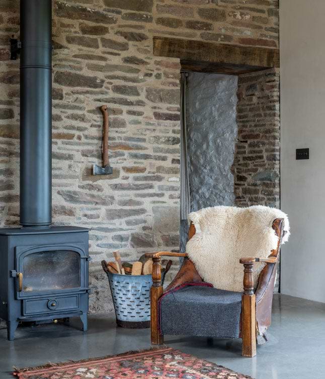 Cozy interior featuring a wooden chair with a fur throw, a black wood stove, and a stone wall adorned with an axe.