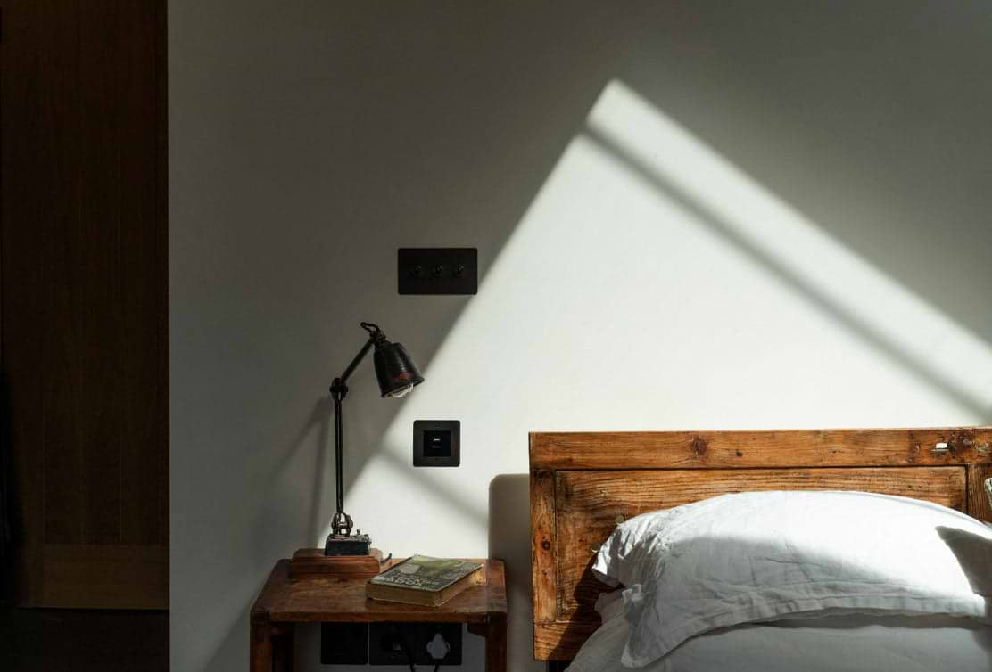A cozy bedroom corner featuring a wooden headboard, side table with a lamp and book, illuminated by soft, diagonal sunlight.