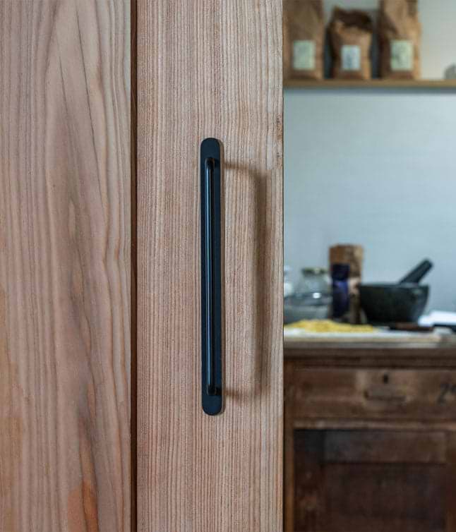 Close-up of a black handle on a wooden door, with a blurred kitchen background featuring rustic decor and cooking ingredients.