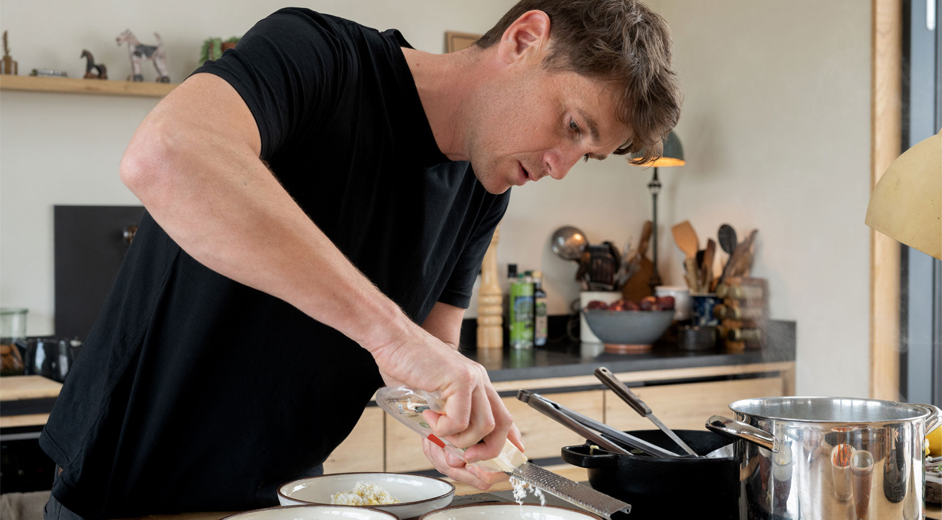 A person prepares food in a modern kitchen, surrounded by cookware, fresh ingredients, and a bowl of pasta.
