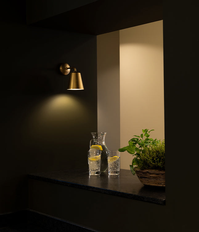A stylish nook featuring a brass wall sconce, two glasses of water with lemon, a pitcher, and a potted plant in a woven basket.