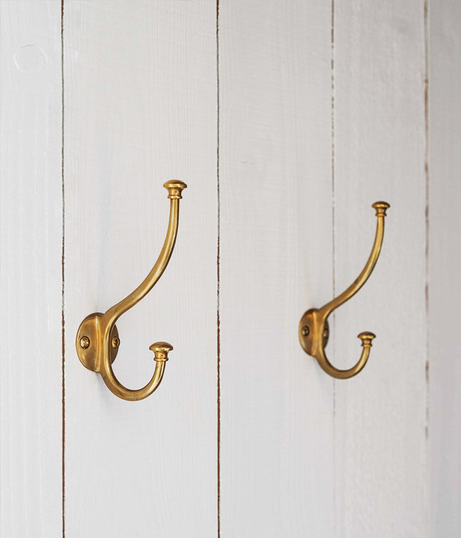 Two elegant brass hooks mounted on a white wooden wall, showcasing a vintage design for hanging items.