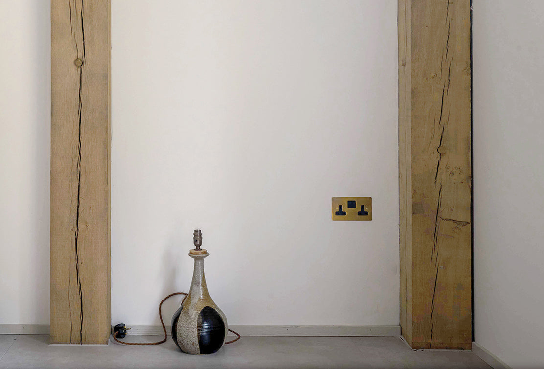 A rustic corner featuring a textured wooden beam, a minimalist ceramic lamp, and a brass electrical outlet against a plain wall.