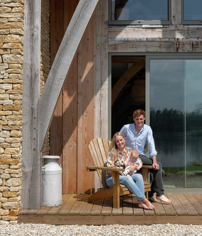 A family enjoys a sunny day on a wooden deck, posing with a baby, with a scenic lakeside view and rustic architecture in the background.