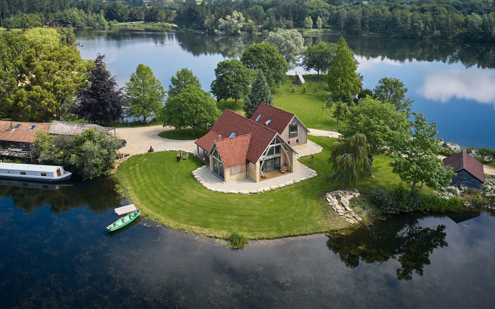 Aerial view of a modern lake house surrounded by lush greenery, with a serene lake reflecting the sky in the background.
