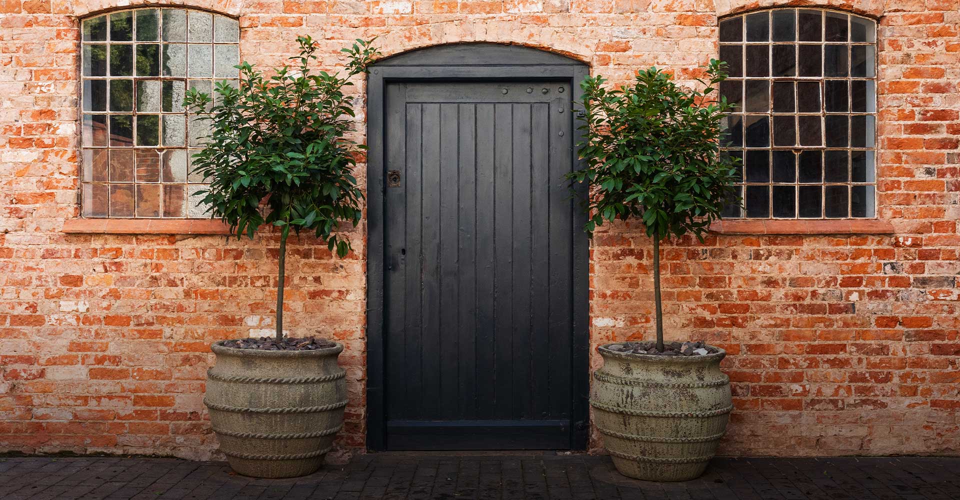 A black door framed by two potted trees stands against a rustic brick wall with large windows, inviting exploration.
