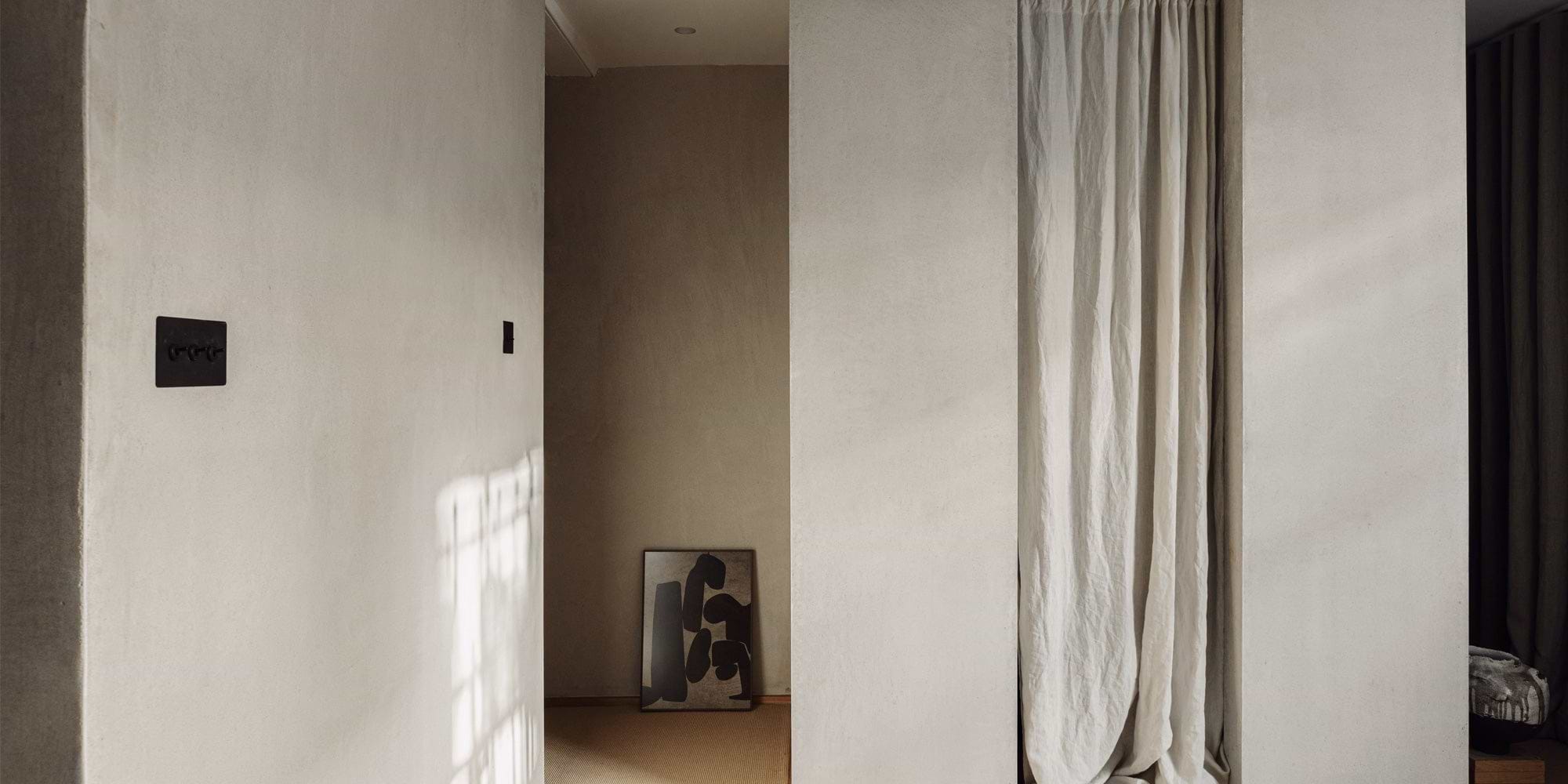 A neutral-toned hallway features a textured wall, a black power switch, and an abstract black artwork against a soft beige background.