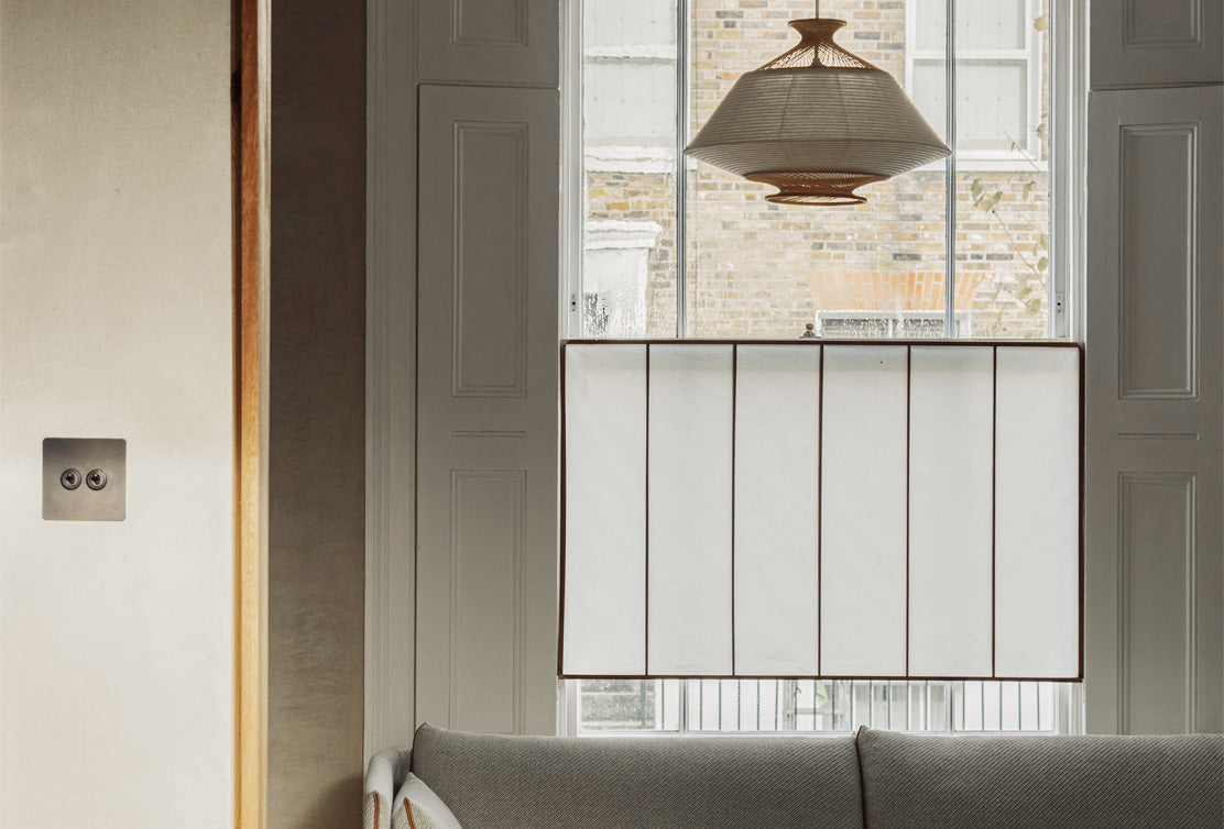A cozy living space featuring a gray sofa, a woven pendant light, and a white window shade, with sunlight filtering through.