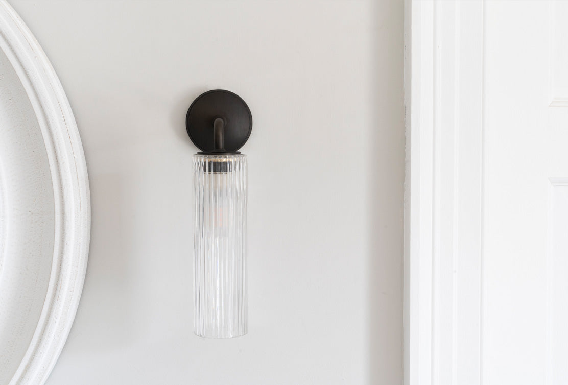 A sleek, modern wall sconce with a clear ribbed glass shade and a bronze metal mount, attached to a light gray wall.