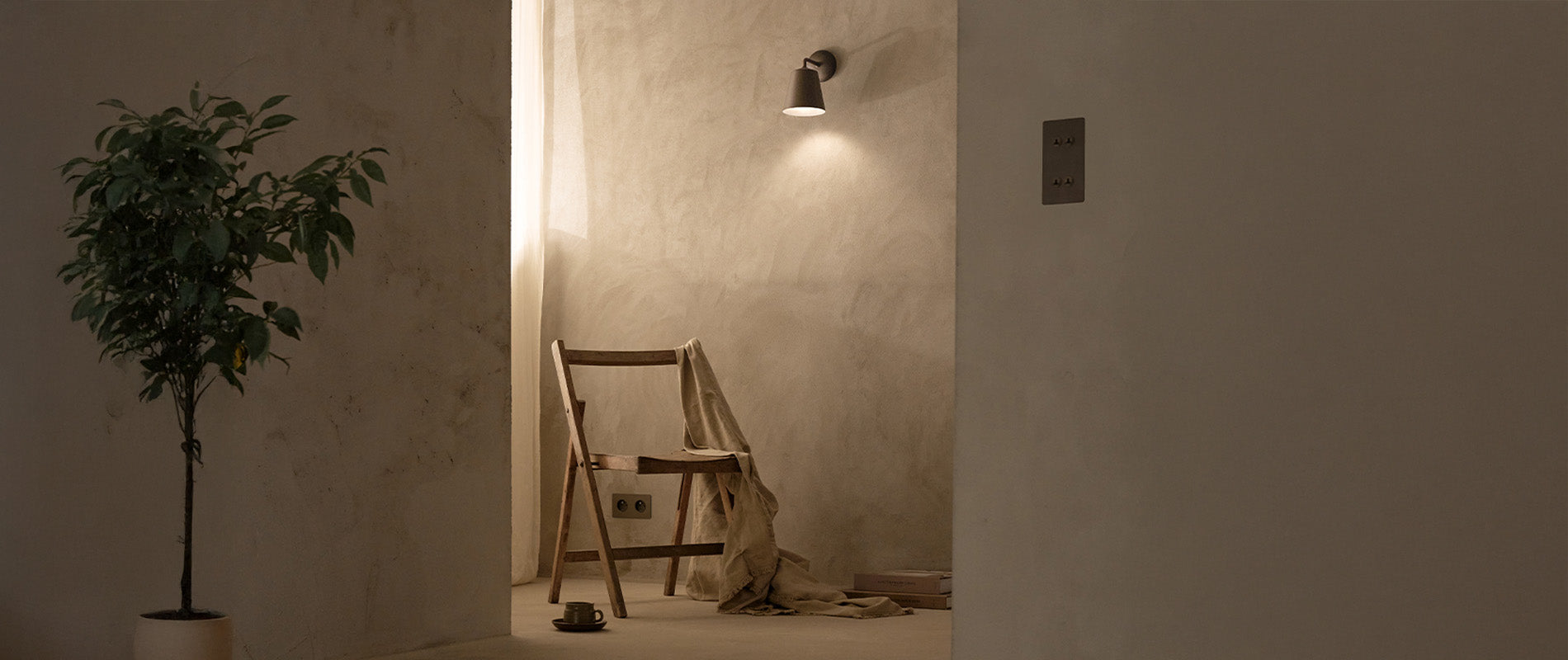 Minimalist room with a wooden chair draped in cloth, a potted plant, and a 'Richmond Wall Light' illuminating the space.
