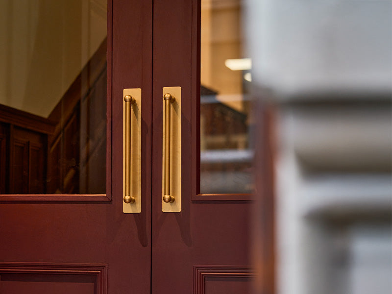 Close-up view of elegant dark red double doors with sleek gold handles and glass panels, revealing a glimpse of interior stairs.
