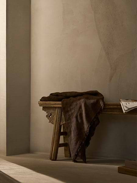 Wooden bench against a limewash wall with a brown blanket and newspaper draped over it.