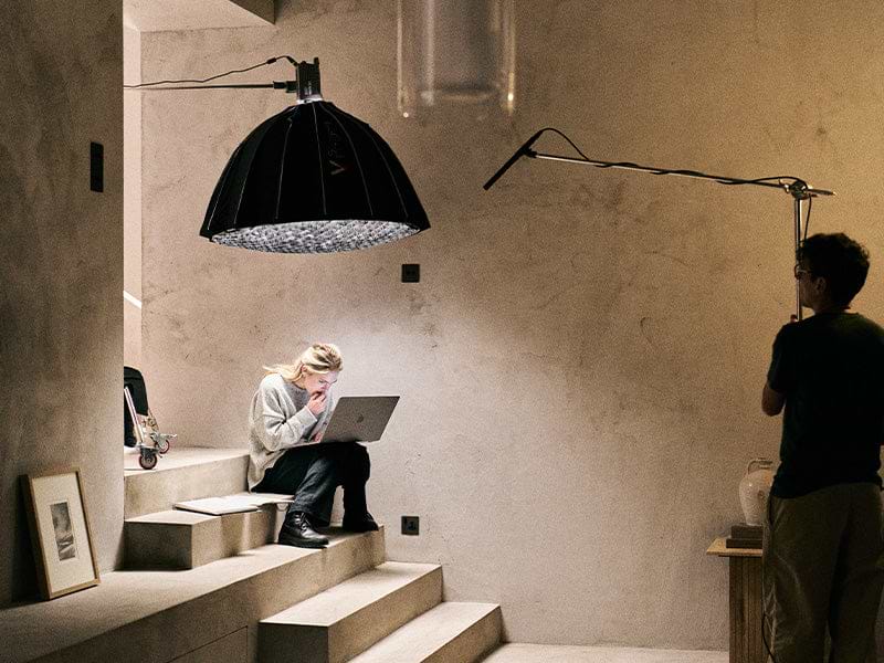 Person sitting on stairs using a laptop under an overhead lamp, another individual stands nearby.
