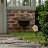Ealing Grond Spotlight in Antiek Messing afwerking op een stenen pad, ideaal voor accentverlichting buitenshuis in de buurt van bakstenen en groen.