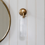 Claremont Kleine Wandlamp met geribbeld glas en antieke messing afwerking tegen een witte lambrisering, naast een ronde spiegel.