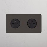 2x Rond Belgisch Type E Stopcontact in brons (zwart inzetstuk), Dubbele Afdekplaat, op witte wand.