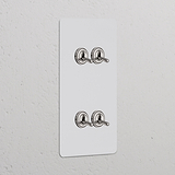 4x Wissel-Tuimelschakelaar (Dubbele Verticale Afdekplaat) in Beschilderbaar Gepolijst Nikkel op een witte muur.