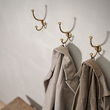 Mews Haak - Antiek Messing: Drie haken aan een lichtgrijze muur om jassen aan op te hangen.
