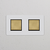 Interrupteurs doubles carrés en finition laiton antique avec inserts noirs sur une plaque murale blanche.