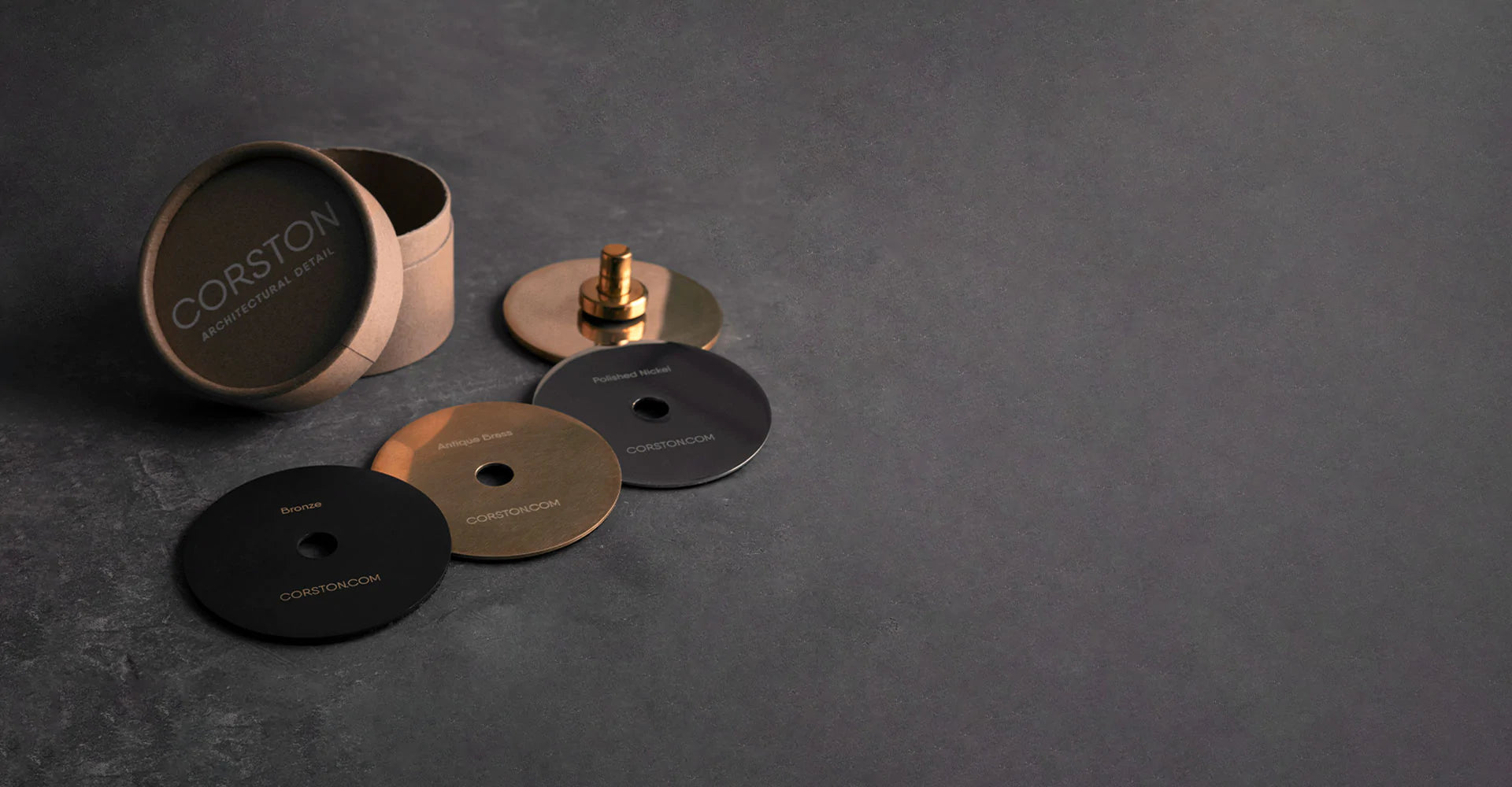 Drei Musterscheiben von Corston (Bronze, Antikes Messing und Poliertes Nickel) neben einer runden Kartonverpackung auf einer grauen Unterlage
