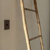 Kipphebelschalter – Bronze – an eine cremefarbene Wand montiert, Leiter aus Bambus rechts.