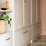 Tirador para mueble Sicomoro de 128 mm en Latón antiguo sobre mueble blanco con planta y jarrón cerca.