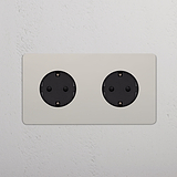 Bases Schuko x2 de doble marco, níquel pulido con insertos negros, montadas sobre pared texturizada blanca.