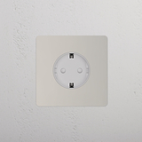 Una toma Schuko de níquel pulido (marco simple) con inserto blanco sobre pared blanca texturizada.