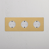 Base triple con módulo Schuko x3 redondo en latón antiguo, inserto blanco, dentro de un marco triple sobre pared blanca.