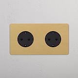 Base de enchufe Schuko doble en marco doble, de latón antiguo con inserto negro, montada en pared blanca.
