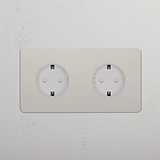 Módulo de doble enchufe Schuko (marco doble) en níquel pulido con inserto blanco sobre pared texturizada blanca.