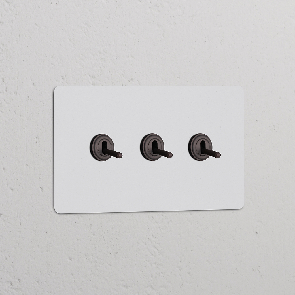 Interruptor conmutador de palanca x3 en marco doble pintable y bronce sobre pared blanca, elegante y funcional.